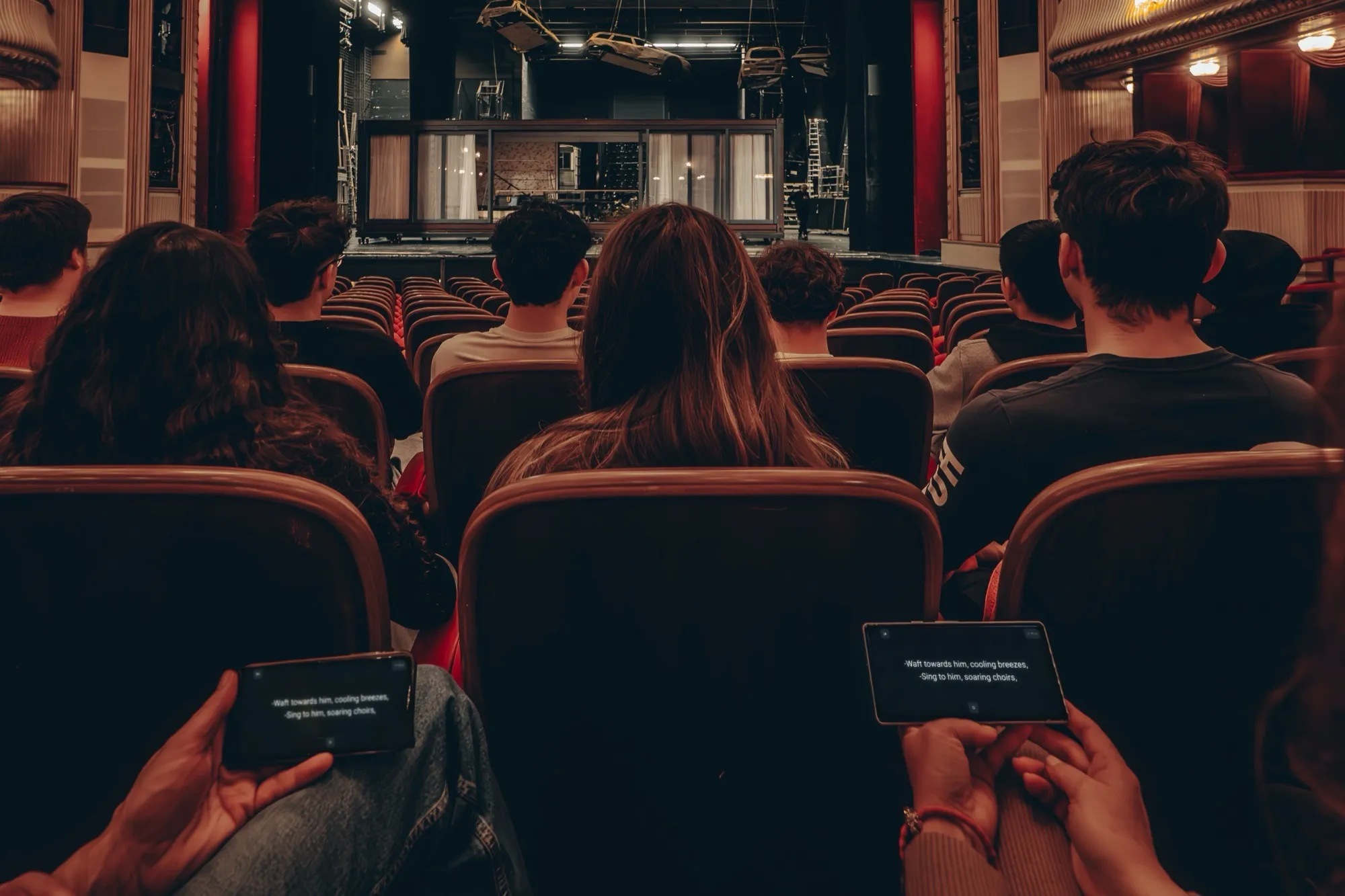 An audience photographed from behind, two people in front holding smartphones sideways with subtitles on their screens
