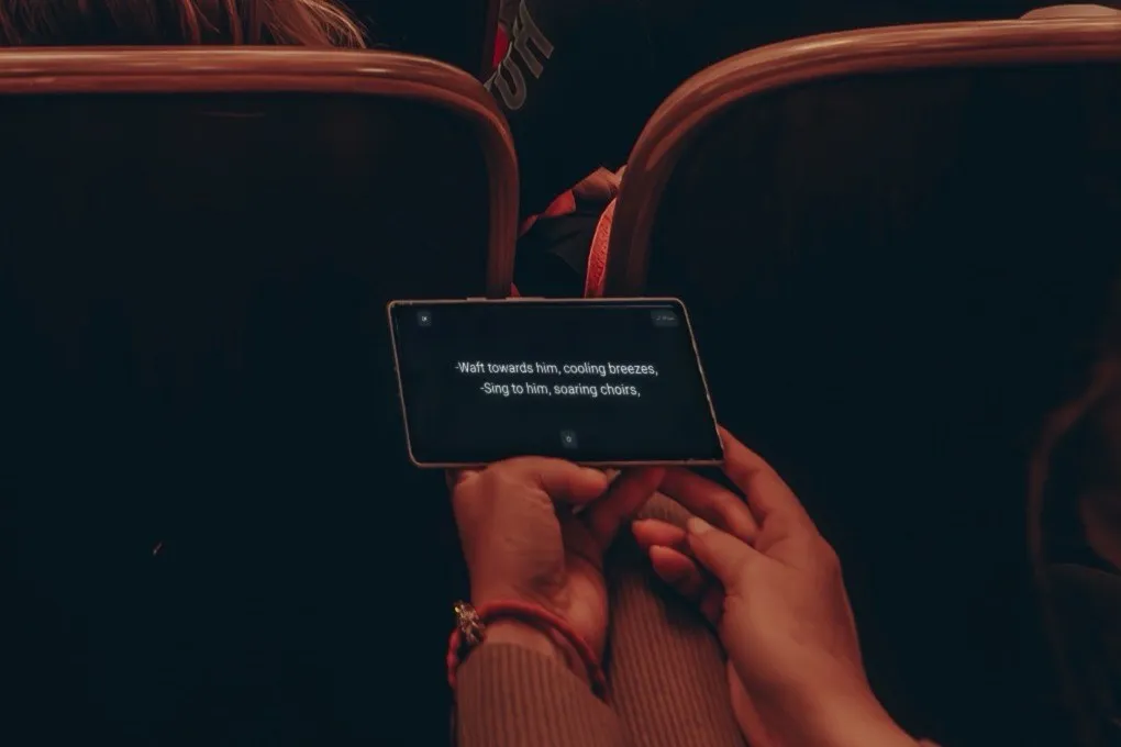 In a dark auditorium, a guest holds a smartphone sideways; the libretto42 app shows subtitles