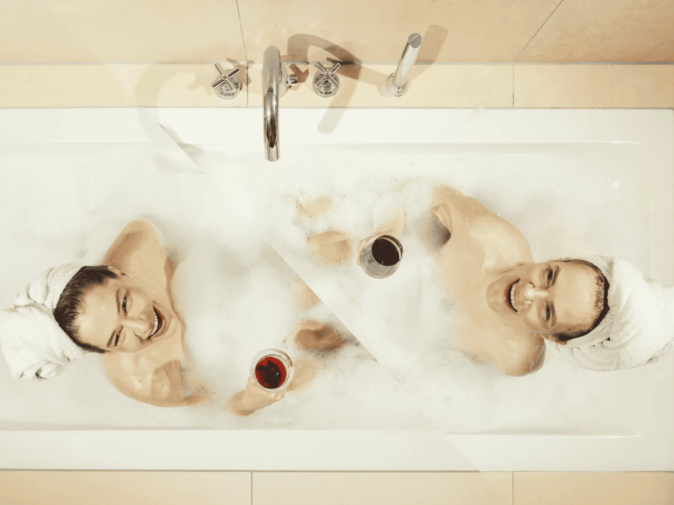 Zwei Menschen mit Handtuch-Turban liegen lachend in einer Schaumbadewanne und halten je ein Glas Rotwein, von oben fotografiert