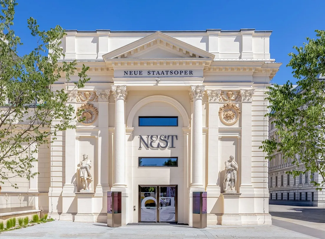 The pale columned façade of the Künstlerhaus with the words Neue Staatsoper and the NEST logo above the entrance, under a blue sky